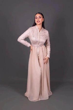 Mannequin portant un caftan marocain beige poudré à manches longues, orné de broderies raffinées sur le buste et d’une jupe fluide longue.