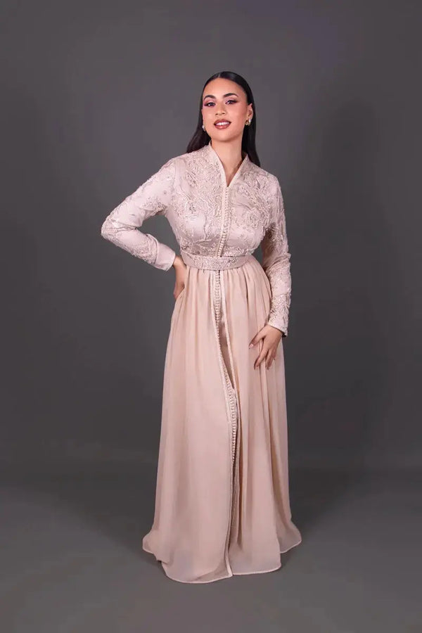 Mannequin portant un caftan marocain beige poudré à manches longues, orné de broderies raffinées sur le buste et d’une jupe fluide longue.
