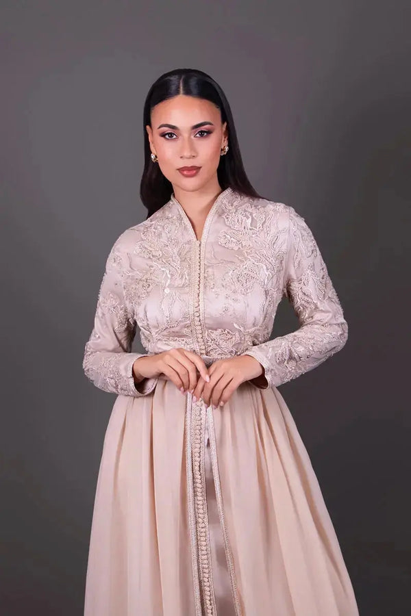 Zoom sur le buste d’un caftan beige à broderies sophistiquées et finitions artisanales, mettant en valeur les détails du tissu et de la coupe.