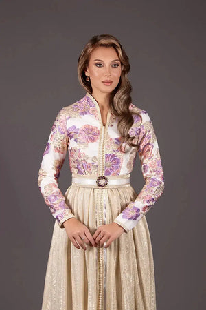 caftan élégant avec un haut brocard  orné de motifs floraux violets et dorés. Elle se tient de face, les mains croisées sur une jupe dorée texturée avec une ceinture bijou.