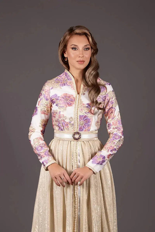 caftan élégant avec un haut brocard  orné de motifs floraux violets et dorés. Elle se tient de face, les mains croisées sur une jupe dorée texturée avec une ceinture bijou.