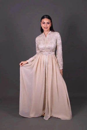 Elegante caftano marocchino beige chiaro con ricami delicati e cintura coordinata, indossato da un manichino che valorizza l’ampiezza della gonna.